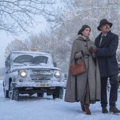 Eine Frau und ein Mann gehen über eine verschneite Straße. Hinter ihnen ist ein Auto zu sehen.
