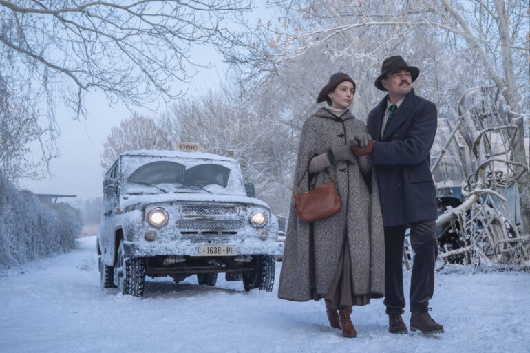 Eine Frau und ein Mann gehen über eine verschneite Straße. Hinter ihnen ist ein Auto zu sehen.