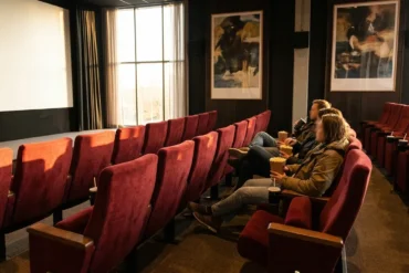 Fotorealistische Szene eines Kinosaals mit rotem Samt, großem Bildschirm und warmem, natürlichem Licht, auf dem mehrere Filmplakate ohne Text als visuelle Motive zu erkennen sind. Drei Personen sitzen entspannt in den mittleren Reihen, Popcorn und Getränke in den Händen, realistische Stoff- und Materialtexturen, cineastische Stimmung ohne Schrift oder Symbole.