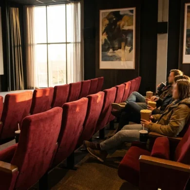 Fotorealistische Szene eines Kinosaals mit rotem Samt, großem Bildschirm und warmem, natürlichem Licht, auf dem mehrere Filmplakate ohne Text als visuelle Motive zu erkennen sind. Drei Personen sitzen entspannt in den mittleren Reihen, Popcorn und Getränke in den Händen, realistische Stoff- und Materialtexturen, cineastische Stimmung ohne Schrift oder Symbole.