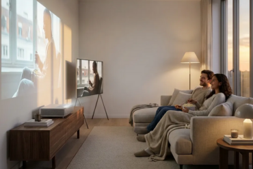 Fotorealistische Szene eines modernen kleinen Wohnzimmers, in dem ein stilvoller Heimkino-Beamer auf einem Lowboard steht und eine große Projektion an eine saubere Wand wirft, daneben ein schlanker großer Fernseher als Vergleich, warmes natürliches Abendlicht durch ein Seitenfenster, realistische Holz-, Stoff- und Metalltexturen. Die Komposition soll aufgeräumt und wohnlich wirken, mit 2 Personen auf einem Sofa in entspannter Filmabend-Stimmung, dezente neutrale Farben, feine Lichtreflexe, keine Schrift, keine Zahlen, no text