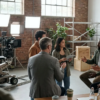 Fotorealistische Szene in einer modernen Filmstudiohalle mit genau 5 Personen an einem nachhaltigen Set: eine Kamera auf Dolly, LED-Scheinwerfer, wiederverwendbare Kulissenelemente aus Holz und Metall, Mehrwegbecher auf einem Produktionstisch, Kabel ordentlich verlegt, natürliches Tageslicht durch hohe Fenster. Ruhige professionelle Stimmung, natürliche Grün-, Grau- und Holztöne, realistische Texturen von Stoff, Aluminium, Holz und Beton, leichte Tiefenschärfe, saubere kinonahe Komposition ohne sichtbare Schrift oder Symbole.