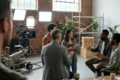 Fotorealistische Szene in einer modernen Filmstudiohalle mit genau 5 Personen an einem nachhaltigen Set: eine Kamera auf Dolly, LED-Scheinwerfer, wiederverwendbare Kulissenelemente aus Holz und Metall, Mehrwegbecher auf einem Produktionstisch, Kabel ordentlich verlegt, natürliches Tageslicht durch hohe Fenster. Ruhige professionelle Stimmung, natürliche Grün-, Grau- und Holztöne, realistische Texturen von Stoff, Aluminium, Holz und Beton, leichte Tiefenschärfe, saubere kinonahe Komposition ohne sichtbare Schrift oder Symbole.