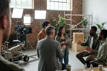 Fotorealistische Szene in einer modernen Filmstudiohalle mit genau 5 Personen an einem nachhaltigen Set: eine Kamera auf Dolly, LED-Scheinwerfer, wiederverwendbare Kulissenelemente aus Holz und Metall, Mehrwegbecher auf einem Produktionstisch, Kabel ordentlich verlegt, natürliches Tageslicht durch hohe Fenster. Ruhige professionelle Stimmung, natürliche Grün-, Grau- und Holztöne, realistische Texturen von Stoff, Aluminium, Holz und Beton, leichte Tiefenschärfe, saubere kinonahe Komposition ohne sichtbare Schrift oder Symbole.