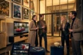 Fotorealistische Szene eines modernen deutschen Kinoabends mit genau 4 Personen in einem stilvollen Foyer, eine große Filmplakatwand ohne lesbare Schrift nur als Lichtfläche im Hintergrund, natürliches warmes Abendlicht durch Glasfronten, realistische Stoff- und Metalltexturen, Popcorn und Kinotickets als dezente Requisiten. Im Vordergrund liegen zusätzlich ein Tablet und ein Fernseher in einem angrenzenden Wohnbereich als Hinweis auf Streaming, harmonische Farben in Rot, Gold und Dunkelblau, scharfe Details, cineastische Stimmung, no text