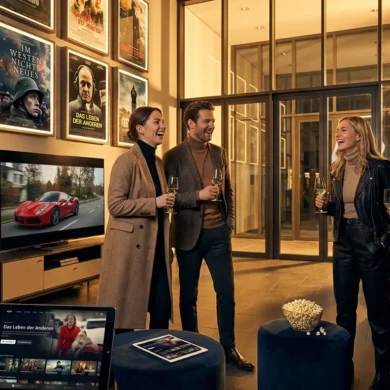 Fotorealistische Szene eines modernen deutschen Kinoabends mit genau 4 Personen in einem stilvollen Foyer, eine große Filmplakatwand ohne lesbare Schrift nur als Lichtfläche im Hintergrund, natürliches warmes Abendlicht durch Glasfronten, realistische Stoff- und Metalltexturen, Popcorn und Kinotickets als dezente Requisiten. Im Vordergrund liegen zusätzlich ein Tablet und ein Fernseher in einem angrenzenden Wohnbereich als Hinweis auf Streaming, harmonische Farben in Rot, Gold und Dunkelblau, scharfe Details, cineastische Stimmung, no text