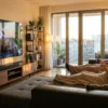 Fotorealistische Szene eines modernen deutschen Wohnzimmers mit echtem Heimkino-Setup, ein großer Fernseher an der Wand, eine hochwertige Dolby-Atmos-Soundbar und zusätzliche Lautsprecher, 2 Personen sitzen entspannt auf einem Sofa, warmes natürliches Abendlicht fällt durch große Fenster und betont realistische Texturen von Holz, Stoff und Metall. Die Komposition wirkt hochwertig, wohnlich und technisch glaubwürdig, mit ruhiger Kino-Stimmung, ausgewogenen dunklen Farben und subtilen Lichtreflexen auf den Geräten, no text.