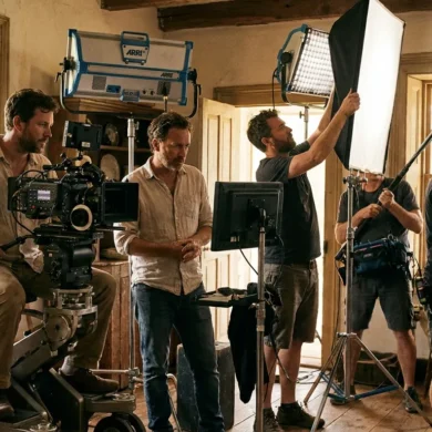 Photorealistische Szene auf einem Filmset während laufender Dreharbeiten, 5 Personen arbeiten konzentriert an Kamera, Licht und Regie in einer realistischen Innenraumkulisse, warme natürliche Beleuchtung mischt sich mit professionellen Filmscheinwerfern, sichtbare Texturen auf Holz, Stoff, Metall und Haut. Die Komposition soll cineastisch, aber glaubwürdig wirken, mit klarer Tiefenwirkung, ruhiger Farbpalette und authentischer Arbeitsatmosphäre, ohne Schrift oder grafische Elemente.