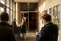 Fotorealistische Szene in einem modernen deutschen Kinofoyer mit 4 Personen, davon eine Person mit Kopfhörern und eine Person mit Smartphone zur Audiodeskription, warmes natürliches Licht aus seitlichen Fensterflächen, realistische Stoffe, Metalloberflächen und Hauttexturen, ruhige einladende Stimmung. Im Hintergrund ein diskreter Blick in den Kinosaal mit roten Sitzen und weichem Licht, ausgewogene Komposition, natürliche Farben, kein Text.