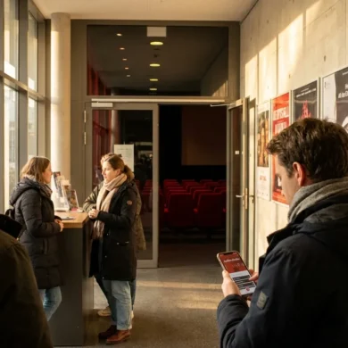 Fotorealistische Szene in einem modernen deutschen Kinofoyer mit 4 Personen, davon eine Person mit Kopfhörern und eine Person mit Smartphone zur Audiodeskription, warmes natürliches Licht aus seitlichen Fensterflächen, realistische Stoffe, Metalloberflächen und Hauttexturen, ruhige einladende Stimmung. Im Hintergrund ein diskreter Blick in den Kinosaal mit roten Sitzen und weichem Licht, ausgewogene Komposition, natürliche Farben, kein Text.