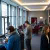 Fotorealistische Szene in einem modernen deutschen Kinofoyer mit genau 4 Personen, darunter eine Person mit Kopfhörern und Smartphone für Audiodeskription und eine Person, die barrierefreie Untertitel auf einem Tablet auswählt. Natürliches Licht fällt durch große Glasflächen, dazu warme Innenbeleuchtung, realistische Texturen von Stoff, Glas, Metall und Haut, ruhige inklusive Stimmung in dezenten Blau-, Grau- und Rottönen, kein sichtbarer Text oder Schrift im Bild, no text