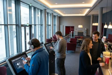 Fotorealistische Szene in einem modernen deutschen Kinofoyer mit genau 4 Personen, darunter eine Person mit Kopfhörern und Smartphone für Audiodeskription und eine Person, die barrierefreie Untertitel auf einem Tablet auswählt. Natürliches Licht fällt durch große Glasflächen, dazu warme Innenbeleuchtung, realistische Texturen von Stoff, Glas, Metall und Haut, ruhige inklusive Stimmung in dezenten Blau-, Grau- und Rottönen, kein sichtbarer Text oder Schrift im Bild, no text