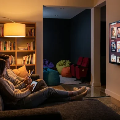Ein fotorealistisches Wohnzimmer am Abend mit genau 2 Personen auf einem Sofa, die auf einem großen Fernseher durch verschiedene Filmgenres und Historienfilme browsen, natürliches warmes Licht von einer Stehlampe und dem Bildschirm, realistische Texturen von Stoff, Holz und Glas. Die Komposition soll modern und aufgeräumt sein, mit gemütlicher Heimkino-Stimmung, klaren Hauttönen, sanften Schatten und sichtbarer Tiefe im Raum, kein Text.