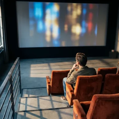 Fotorealistische Szene in einem stilvollen, modernen Kinosaal mit natürlichem Licht, das durch seitliche Fenster in ein Foyer mit Filmplakatrahmen ohne Schrift fällt; im Vordergrund sitzt genau eine einzelne Person in einer roten Kinositzreihe und betrachtet konzentriert eine große Leinwand mit abstrakten filmischen Lichtstimmungen, während im Hintergrund unscharf eine Filmkamera und ein Schnittmonitor zu sehen sind. Warme Rot- und Goldtöne treffen auf kühle Schatten, realistische Stoff- und Metalltexturen, dezente Tiefenschärfe, cineastische Stimmung, hochwertig und professionell, ohne Text, ohne Zahlen, ohne Logos, ohne Schriftzeichen.
