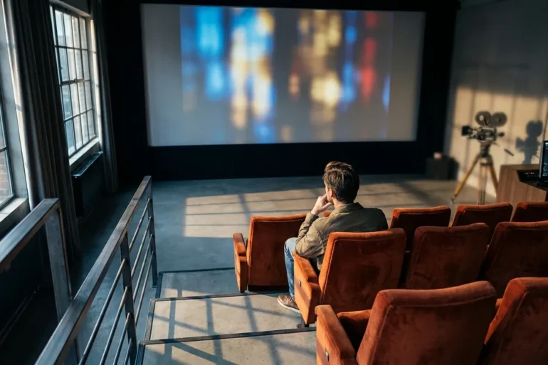 Fotorealistische Szene in einem stilvollen, modernen Kinosaal mit natürlichem Licht, das durch seitliche Fenster in ein Foyer mit Filmplakatrahmen ohne Schrift fällt; im Vordergrund sitzt genau eine einzelne Person in einer roten Kinositzreihe und betrachtet konzentriert eine große Leinwand mit abstrakten filmischen Lichtstimmungen, während im Hintergrund unscharf eine Filmkamera und ein Schnittmonitor zu sehen sind. Warme Rot- und Goldtöne treffen auf kühle Schatten, realistische Stoff- und Metalltexturen, dezente Tiefenschärfe, cineastische Stimmung, hochwertig und professionell, ohne Text, ohne Zahlen, ohne Logos, ohne Schriftzeichen.