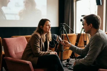 Photorealistische Szene eines intimen Regie-Interviews in einem modernen deutschen Kino: eine einzelne Regisseurin sitzt leicht seitlich vor der Leinwand, gegenüber ein Interviewer mit kleinem Mikrofon, im Hintergrund unscharf Kameratechnik und leere Kinositze. Natürliches warmes Licht, realistische Haut- und Stofftexturen, ruhige cineastische Stimmung in gedeckten Rot- und Brauntönen, keine Schrift, keine Logos, no text