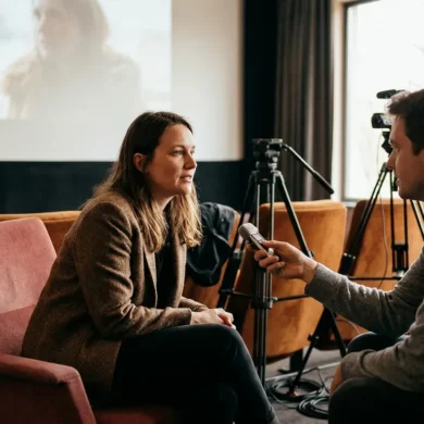 Photorealistische Szene eines intimen Regie-Interviews in einem modernen deutschen Kino: eine einzelne Regisseurin sitzt leicht seitlich vor der Leinwand, gegenüber ein Interviewer mit kleinem Mikrofon, im Hintergrund unscharf Kameratechnik und leere Kinositze. Natürliches warmes Licht, realistische Haut- und Stofftexturen, ruhige cineastische Stimmung in gedeckten Rot- und Brauntönen, keine Schrift, keine Logos, no text