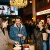 Fotorealistische Szene in einem modernen deutschen Kinofoyer mit genau 4 Personen, die vor einer beleuchteten Ticket- und Snackzone ihre Auswahl treffen, natürliches warmes Abendlicht mischt sich mit weichem Innenlicht, realistische Texturen von Stoff, Glas, Metall und Papier. Fokus auf die Stimmung eines geplanten Kinobesuchs mit sichtbarer Premium-Atmosphäre, elegante Komposition mit Tiefenschärfe, natürliche Farben in Rot, Dunkelblau und Gold, keine Schrift, keine Zahlen, kein Text.