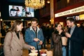 Fotorealistische Szene in einem modernen deutschen Kinofoyer mit genau 4 Personen, die vor einer beleuchteten Ticket- und Snackzone ihre Auswahl treffen, natürliches warmes Abendlicht mischt sich mit weichem Innenlicht, realistische Texturen von Stoff, Glas, Metall und Papier. Fokus auf die Stimmung eines geplanten Kinobesuchs mit sichtbarer Premium-Atmosphäre, elegante Komposition mit Tiefenschärfe, natürliche Farben in Rot, Dunkelblau und Gold, keine Schrift, keine Zahlen, kein Text.