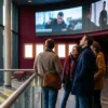 Fotorealistische Szene in einem modernen Kinofoyer in Deutschland, 3 Personen betrachten aufmerksam eine große digitale Trailer-Vorschau an einer Wand ohne lesbare Schrift, warmes natürliches Licht durch Glasfronten, realistische Texturen auf Stoffen, Haut und Metall, elegante cineastische Stimmung. Im Hintergrund dezente Kinokassen und Posterflächen ohne Text, ausgewogene Komposition mit Tiefenschärfe, natürliche Farben in Rot, Dunkelblau und Gold, no text