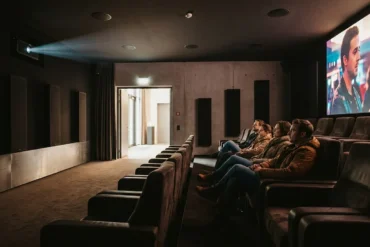 Fotorealistische Szene in einem modernen deutschen Kino mit genau 4 Personen, die in bequemen Sesseln sitzen und konzentriert auf eine große Leinwand blicken, während dezente Deckenlautsprecher und seitliche Lautsprecher die Dolby-Atmos-Umgebung sichtbar machen, natürliches Licht nur sanft aus dem Foyer einfallend, realistische Stoff-, Leder- und Metalltexturen, warme dunkle Farbstimmung mit cineastischem Kontrast. Fokus auf immersives Premium-Kinoerlebnis, klare Tiefenwirkung im Saal, authentische Architektur eines hochwertigen Kinosaals, keine Schrift, keine Logos, keine Zahlen, keine sonstigen Textelemente im Bild.