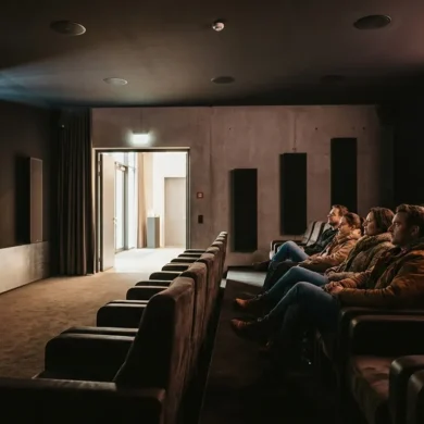 Fotorealistische Szene in einem modernen deutschen Kino mit genau 4 Personen, die in bequemen Sesseln sitzen und konzentriert auf eine große Leinwand blicken, während dezente Deckenlautsprecher und seitliche Lautsprecher die Dolby-Atmos-Umgebung sichtbar machen, natürliches Licht nur sanft aus dem Foyer einfallend, realistische Stoff-, Leder- und Metalltexturen, warme dunkle Farbstimmung mit cineastischem Kontrast. Fokus auf immersives Premium-Kinoerlebnis, klare Tiefenwirkung im Saal, authentische Architektur eines hochwertigen Kinosaals, keine Schrift, keine Logos, keine Zahlen, keine sonstigen Textelemente im Bild.