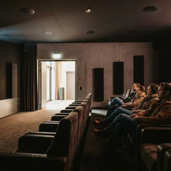 Fotorealistische Szene in einem modernen deutschen Kino mit genau 4 Personen, die in bequemen Sesseln sitzen und konzentriert auf eine große Leinwand blicken, während dezente Deckenlautsprecher und seitliche Lautsprecher die Dolby-Atmos-Umgebung sichtbar machen, natürliches Licht nur sanft aus dem Foyer einfallend, realistische Stoff-, Leder- und Metalltexturen, warme dunkle Farbstimmung mit cineastischem Kontrast. Fokus auf immersives Premium-Kinoerlebnis, klare Tiefenwirkung im Saal, authentische Architektur eines hochwertigen Kinosaals, keine Schrift, keine Logos, keine Zahlen, keine sonstigen Textelemente im Bild.