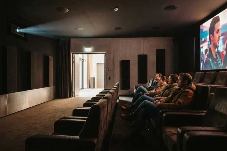 Fotorealistische Szene in einem modernen deutschen Kino mit genau 4 Personen, die in bequemen Sesseln sitzen und konzentriert auf eine große Leinwand blicken, während dezente Deckenlautsprecher und seitliche Lautsprecher die Dolby-Atmos-Umgebung sichtbar machen, natürliches Licht nur sanft aus dem Foyer einfallend, realistische Stoff-, Leder- und Metalltexturen, warme dunkle Farbstimmung mit cineastischem Kontrast. Fokus auf immersives Premium-Kinoerlebnis, klare Tiefenwirkung im Saal, authentische Architektur eines hochwertigen Kinosaals, keine Schrift, keine Logos, keine Zahlen, keine sonstigen Textelemente im Bild.