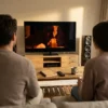Fotorealistische Szene in einem modernen, gemütlichen Wohnzimmer mit natürlichem Fensterlicht: 2 Personen sitzen vor einem großen Fernseher, auf dem eine filmische Szene mit deutlicher Lichtstimmung, Nahaufnahme und starken Farbkontrasten zu sehen ist, daneben liegen Notizbuch und Fernbedienung auf einem Holztisch, realistische Texturen von Stoff, Holz und Glas. Warme, natürliche Farbtöne im Raum, klare Tiefenschärfe, ruhige cineastische Komposition, professioneller Editorial-Look ohne Text, ohne Zahlen, ohne Schrift im Bild.