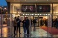 Fotorealistische Szene in natürlichem Licht vor einem modernen Arthouse-Kino in Berlin am frühen Abend, 4 Personen stehen vor Filmplakatrahmen ohne sichtbare Schrift und diskutieren angeregt, warme Reflexionen auf nassem Pflaster, reale Hauttexturen, Stoffe, Glas und Beton sehr detailreich. Im Hintergrund dezente Festival-Atmosphäre mit rotem Teppich ohne Logos oder Text, cineastische Tiefenschärfe, natürliche Farben zwischen warmem Gold, kühlem Blau und urbanem Grau, Stimmung neugierig, kulturell und inspirierend.