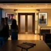 Fotorealistische Fotografie einer modernen Kinohalle vor Beginn einer Preview, mit genau 4 Personen im Vordergrund, die auf eine beleuchtete Saaltür und Filmplakate ohne lesbare Schrift blicken, natürliches warmes Abendlicht aus dem Foyer, realistische Texturen von Samtsitzen, Glas, Metall und Papier. Stimmung gespannt und einladend, cineastische Komposition mit klarer Tiefenschärfe, dezente Rot- und Goldtöne, professioneller Look für einen Artikel über Kino Preview und Kinoevent, keinerlei Text oder Schrift im Bild.