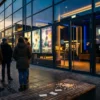 Fotorealistische Szene vor einem modernen Kino am frühen Abend, 5 Personen stehen vor leuchtenden Filmplakaten ohne lesbare Schrift und schauen gespannt auf die Eingangsfront, warmes natürliches Licht, realistische Spiegelungen auf Glas, feine Stoff- und Hauttexturen, cineastische Atmosphäre. Im Vordergrund liegen Popcorn und Eintrittskarten ohne Text auf einer Bank, im Hintergrund dezente Stadtlichter, ausgewogene Komposition in goldenen und tiefblauen Farbtönen, no text