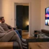 Fotorealistische Szene in einem modernen deutschen Wohnzimmer bei natürlichem Abendlicht: 3 Personen sitzen entspannt vor einem großen Fernseher mit filmischer Beleuchtung, auf dem farbintensive, aber textfreie Streaming-Oberflächen und cineastische Vorschaubilder ohne Schrift zu sehen sind; im Raum stehen ein Sofa, eine Soundbar, ein Beistelltisch mit Snacks und dezente Heimkino-Technik mit realistischen Materialien wie Stoff, Holz und Glas. Warme, natürliche Farbtöne, weiche Schatten, klare Texturen und eine ruhige, hochwertige Stimmung, die den Wandel der Streamingdienste, neue Filme und Filmtrends 2026 visuell vermittelt, ohne jegliche Schrift, Zahlen oder Logos im Bild.
