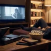 Ein fotorealistisches Wohnzimmer-Heimkino bei natürlichem Abendlicht, eine einzelne Person sitzt entspannt vor einem großen Fernseher, daneben ein geöffneter Blu-ray-Player und mehrere hochwertige Disc-Hüllen auf einem Holztisch, realistische Texturen von Metall, Kunststoff und Papier, warme dezente Farben und ruhige cineastische Stimmung. Im Hintergrund ein ordentliches Regal mit Filmen, weiche Schatten, glaubwürdige Wohnatmosphäre, moderner aber gemütlicher Look, Fokus auf Heimkino, Sammelleidenschaft und hochwertige Unterhaltung ohne sichtbare Schrift oder Symbole.