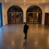 Fotorealistische Szene in einem modernen deutschen Kinofoyer bei natürlichem Abendlicht, eine einzelne Person steht vor beleuchteten Filmplakaten und blickt auf ihr Smartphone, während im Hintergrund unscharf der Eingang zu mehreren Kinosälen sichtbar ist. Warme Gold- und Rottöne, realistische Texturen von Stoff, Glas und Metall, dezente Reflexionen auf dem Boden, glaubwürdige Alltagssituation mit ruhiger, einladender Stimmung und klarem Fokus auf die Entscheidung zwischen Filmstarts im Kino und persönlicher Filmauswahl, ohne jegliche Schrift im Bild.