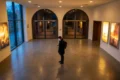 Fotorealistische Szene in einem modernen deutschen Kinofoyer bei natürlichem Abendlicht, eine einzelne Person steht vor beleuchteten Filmplakaten und blickt auf ihr Smartphone, während im Hintergrund unscharf der Eingang zu mehreren Kinosälen sichtbar ist. Warme Gold- und Rottöne, realistische Texturen von Stoff, Glas und Metall, dezente Reflexionen auf dem Boden, glaubwürdige Alltagssituation mit ruhiger, einladender Stimmung und klarem Fokus auf die Entscheidung zwischen Filmstarts im Kino und persönlicher Filmauswahl, ohne jegliche Schrift im Bild.