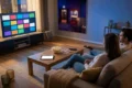 Fotorealistische Szene in einem modernen Wohnzimmer bei natürlicher Abendbeleuchtung: 2 Personen sitzen entspannt auf einem Sofa, vor ihnen ein Couchtisch mit Fernbedienung, Tablet und Popcorn, im Hintergrund ein großer Fernseher mit klar erkennbarer, aber textfreier Streaming-Oberfläche aus farbigen Kacheln ohne Logos oder Schrift. Warme, realistische Texturen bei Stoff, Holz und Glas, dezente Blautöne vom Bildschirmlicht, glaubwürdige Alltagsatmosphäre, hochwertig, aufgeräumt und visuell passend zu einem Vergleich der besten Streamingdienste für Filme, Serien und Sport.