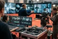 Photorealistische Szene in einem modernen Filmstudio mit genau 3 Personen: ein Regisseur vor mehreren Monitoren, ein Editor an einer Schnittstation und eine Kamerafrau neben professionellem Kamera-Equipment, während auf den Bildschirmen abstrakte visuelle Effekte und Filmsequenzen ohne jegliche Schrift zu sehen sind. Natürliches, weiches Studiolicht, realistische Hauttöne, detaillierte Texturen von Metall, Glas, Kabeln und Stoffen, cineastische Farbgebung in kühlen Blau- und warmen Bernsteintönen, ruhige konzentrierte Stimmung mit hochwertiger, glaubwürdiger Produktionsumgebung.