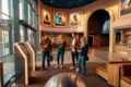 Innenansicht eines modernen Multiplex-Kinos im CinemaxX-Stil mit 4 Personen im Foyer vor dezent beleuchteten Ticket-Terminals, warme natürliche Lichtstimmung durch große Glasfronten, realistische Materialien wie gebürstetes Metall, Teppich, Glas und Leder, klarer Fokus auf Kinoatmosphäre, Wegführung und entspanntes Ankommen. Im Hintergrund sichtbar ein großzügiger Eingangsbereich zu den Sälen und eine subtile Snack-Theke ohne lesbare Beschriftung, fotorealistische Texturen, natürliche Farben, ausgewogene Komposition, keine Schrift, keine Zahlen, keine Logos.