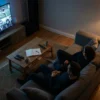 Ein moderner Wohnzimmerabend mit genau 2 Personen auf einem Sofa, vor einem großen Fernseher mit einer spannenden dokumentarischen Bildwelt aus Archivmaterial, Sportmomenten und investigativer Stimmung, dazu verstreute Fernbedienung, Notizbuch und gedimmtes warmes Licht aus einer Stehlampe. Fotorealistische Szene mit natürlicher Beleuchtung, realistischen Stoff- und Holztexturen, kontrastreichen Blautönen und warmen Akzenten, ruhige aber fesselnde Atmosphäre ohne Text, ohne Logos, ohne sichtbare Schriftzeichen.