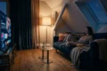 Fotorealistische Szene in einem gemütlichen deutschen Wohnzimmer am Abend: eine einzelne Person sitzt entspannt auf einem Sofa mit Fernbedienung in der Hand, auf dem großen Fernseher ist eine farbenfrohe Streaming-Oberfläche ohne lesbare Schrift zu sehen, daneben liegen Snacks und eine Decke, warmes natürliches Licht von einer Stehlampe trifft auf dezentes Blau aus dem Bildschirm. Realistische Texturen bei Stoffen, Holz und Glas, klare Tiefenschärfe, moderne aber wohnliche Einrichtung, stimmungsvolle Farben zwischen Dunkelblau, Orange und warmen Beigetönen, ein hochwertiger Editorial-Look ohne Text, Zahlen oder Logos im Bild.