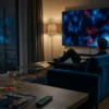 Ein dunkles, stilvolles Wohnzimmer am Abend mit genau 1 Person auf einem Sofa, die konzentriert auf einen großen Fernseher blickt, während auf dem Bildschirm nur abstrakte, spannungsgeladene Lichtstimmungen in Blau, Schwarz und kaltem Rot zu sehen sind, ohne Text oder Logos. Natürliche Lichtquellen durch gedämpfte Stehlampe und schwaches Stadtlicht von draußen, fotorealistische Fotografie mit realistischen Stofftexturen, Hauttönen, Reflexionen auf Glas und Metall, cineastische Komposition mit moderner Streaming-Atmosphäre und subtiler Thriller-Stimmung.